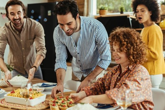 Group of friends laughing and setting food on the table together during a casual home dinner