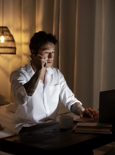 Man working at a laptop late at night while on a phone call, illuminated by warm lamp light — representing consistent effort after hours