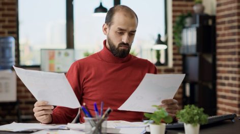 Man comparing two documents in each hand at a cluttered desk, focused and unaware of the camera