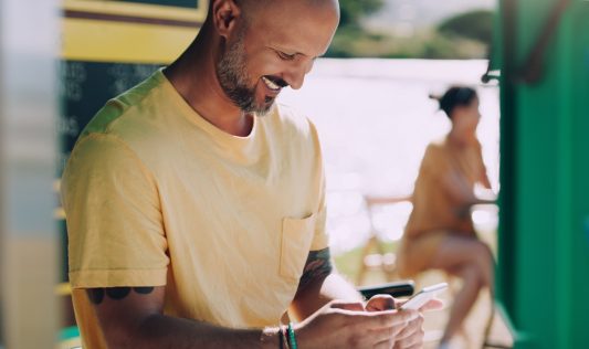 Man smiling warmly while looking at his phone at an outdoor café, another person blurred in the background