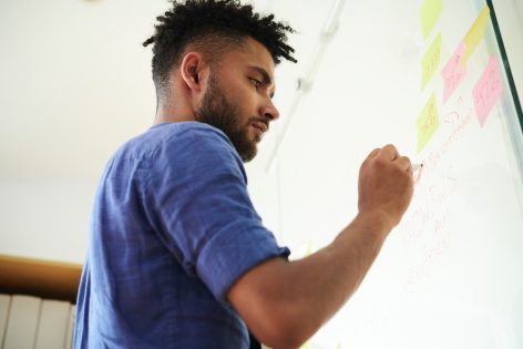 Man writing notes on a glass whiteboard covered in sticky notes — representing a goal that's part of a larger plan