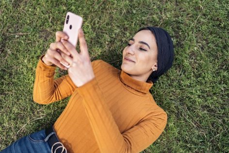 Woman relaxing on the grass, smiling while checking her phone — representing a gentle, welcome nudge rather than a nagging notification